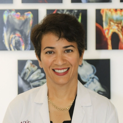 Portrait of Dr. Parisa Gheidarpour wearing a white coat positioned in front of framed dental imagery on a wall.