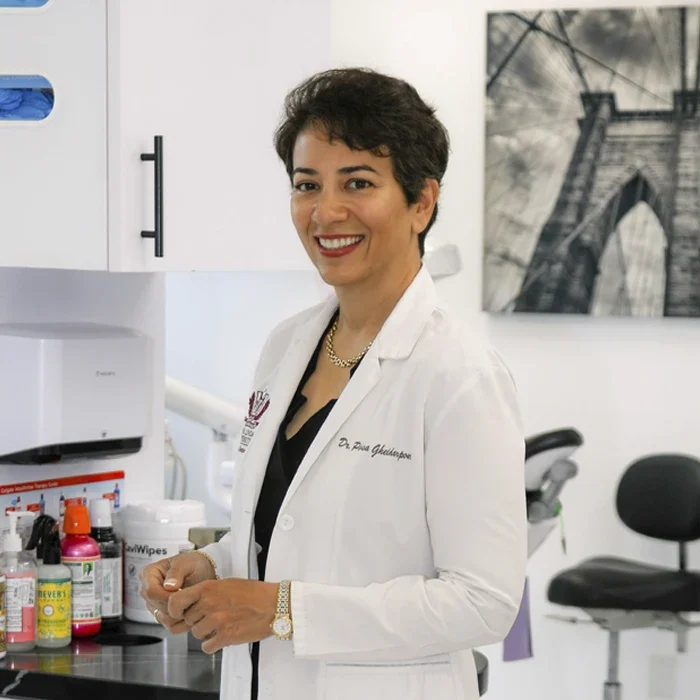 Dr. Parisa Gheidarpour wearing a white coat standing beside dental equipment and cabinetry inside a treatment room.