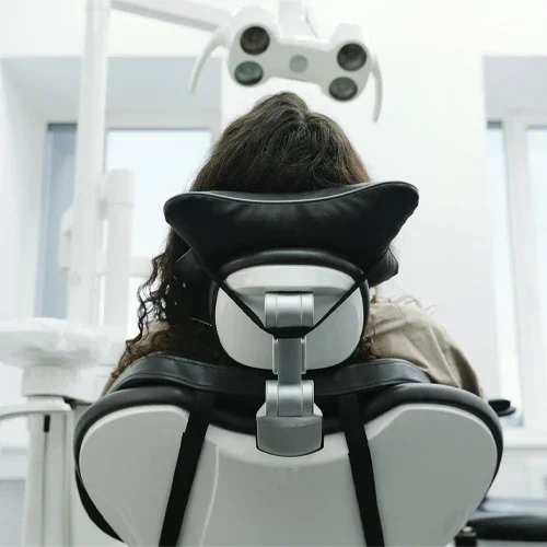 Person seated in a dental chair viewed from behind, with overhead examination equipment visible above.